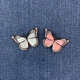 Two butterfly-shaped patches, one pale blue and one pink, with black outlines and white-tipped wings, placed on a blue denim fabric background.