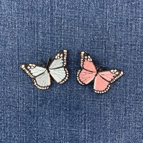 Two butterfly-shaped patches, one pale blue and one pink, with black outlines and white-tipped wings, placed on a blue denim fabric background.