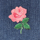 A patch depicting a pink rose with green leaves on a white background, displayed on a denim background