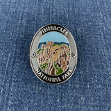 An oval enamel Walking Stick Medallion featuring a landscape illustration of Pinnacles National Park, displayed on a blue denim fabric background.