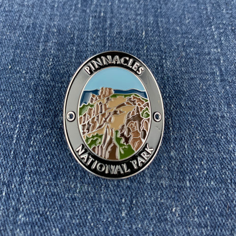 An oval enamel Walking Stick Medallion featuring a landscape illustration of Pinnacles National Park, displayed on a blue denim fabric background.
