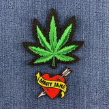 An embroidered patch featuring a green cannabis leaf with a black outline, positioned above a red and yellow 'Mary Jane' heart patch with an arrow, displayed on a blue denim background.