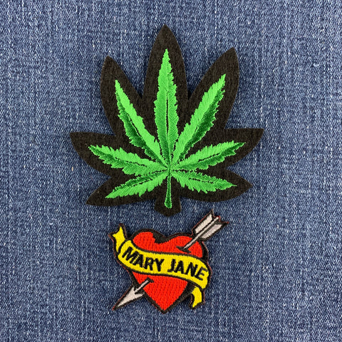 An embroidered patch featuring a green cannabis leaf with a black outline, positioned above a red and yellow 'Mary Jane' heart patch with an arrow, displayed on a blue denim background.