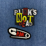 Two embroidered patches displayed on a blue denim fabric background. The top patch features the text 'PUNK'S NOT DEAD' in white, yellow, red, and black. The bottom patch is a safety pin with the word 'PUNK'
