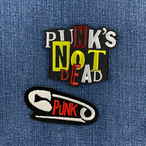 Two embroidered patches displayed on a blue denim fabric background. The top patch features the text 'PUNK'S NOT DEAD' in white, yellow, red, and black. The bottom patch is a safety pin with the word 'PUNK'