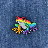 A colorful embroidered frog patch with a rainbow gradient and black stitched outline, placed on a blue denim fabric background.