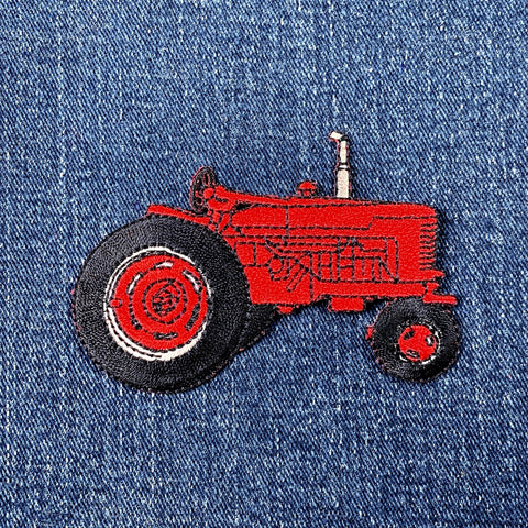 A red tractor applique patch with black wheels and details designed for iron-on application, displayed on a denim background