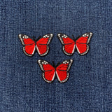 Three identical embroidered patches of a butterfly with bright red wings, black borders, and small white details, arranged on a denim background.