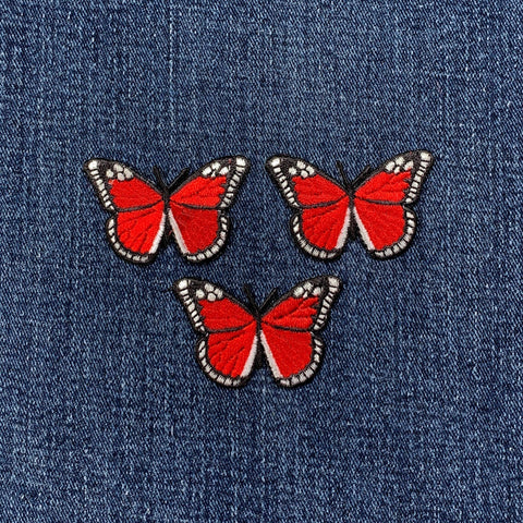 Three identical embroidered patches of a butterfly with bright red wings, black borders, and small white details, arranged on a denim background.