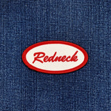 An oval embroidered patch with a red border and white center featuring the word 'Redneck' in red script, shown on a blue denim fabric background.