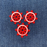 Three identical embroidered patches of a ship's wheel, colored bright red with a white outline around the rim, arranged on a denim background
