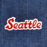 A close-up of a red embroidered 'Seattle' patch with a white border, applied to a blue denim fabric background.