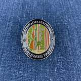 An oval enamel Walking Stick Medallion featuring Redwood National and State Parks, displayed on a blue denim fabric background.
