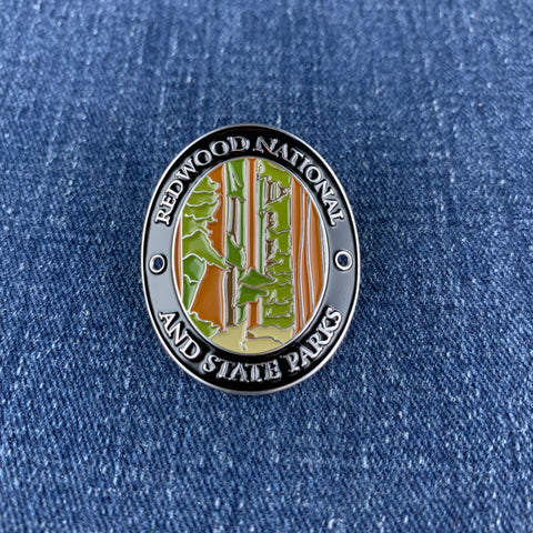 An oval enamel Walking Stick Medallion featuring Redwood National and State Parks, displayed on a blue denim fabric background.