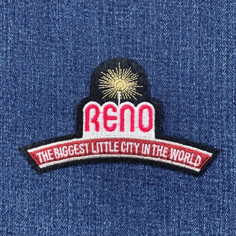 An applique patch with the text 'RENO THE BIGGEST LITTLE CITY IN THE WORLD' on it, predominantly red and black with a gold starburst on a denim background.