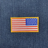 A reverse embroidered American flag patch with a yellow border, displayed on a blue denim fabric background.