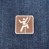 A square brown patch with a white border and a white climbing icon, placed on a blue denim fabric background.