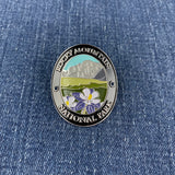 An oval metal Walking Stick Medallion featuring a colorful landscape of Rocky Mountain National Park with a mountain, fields, and purple flowers, displayed on a blue denim background.