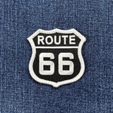 A shield-shaped embroidered patch of the Route 66 highway sign. The patch has a black background with "ROUTE" in white text at the top and "66" in large white numbers below, surrounded by a white border. It is displayed against a denim fabric.