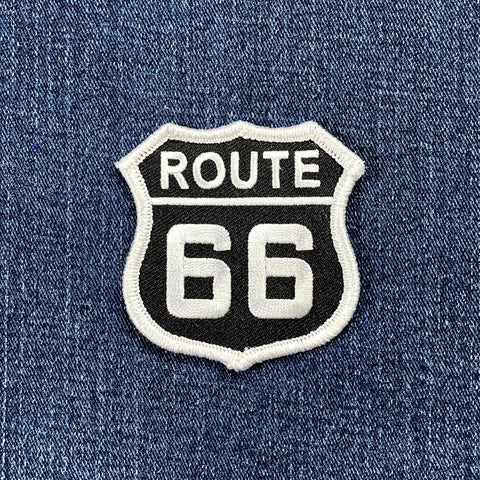 A shield-shaped embroidered patch of the Route 66 highway sign. The patch has a black background with "ROUTE" in white text at the top and "66" in large white numbers below, surrounded by a white border. It is displayed against a denim fabric.