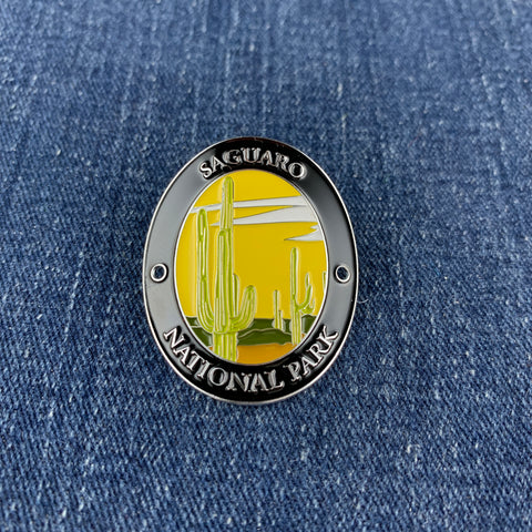 A medallion with an illustration of saguaro cacti and the text 'Saguaro National Park' meant to be affixed to a walking stick, displayed on a denim background