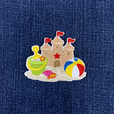 A beach and sandcastle patch which includes a tan sandcastle with red flags, a green bucket, a red star, a pink seashell, and a red, white, blue, and yellow beach ball on a white base.