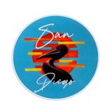 A circular sticker of a pelican is set against a striped orange and yellow sunset on a bright blue background. The words 'San Diego' are written above and below the bird.