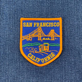 An orange-bordered, shield-shaped embroidered patch on blue denim shows the Golden Gate Bridge, a cable car, and hills.