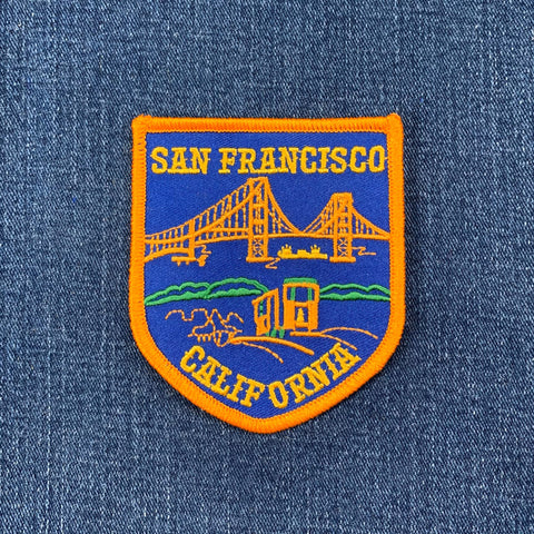 An orange-bordered, shield-shaped embroidered patch on blue denim shows the Golden Gate Bridge, a cable car, and hills.