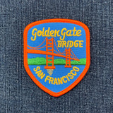 A shield-shaped embroidered patch featuring the Golden Gate Bridge and 'San Francisco' text, placed on a blue denim fabric background.