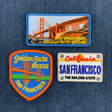 Three San Francisco Golden Gate Bridge themed embroidered patches displayed on a blue denim background. The set includes a rectangular patch with the bridge and 'SAN FRANCISCO GOLDEN GATE BRIDGE' text, a shield-shaped patch with 'Golden Gate Bridge SAN FRANCISCO' on a blue background, and a California license plate style patch reading 'SAN FRANCISCO THE GOLDEN STATE'.