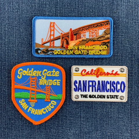 Three San Francisco Golden Gate Bridge themed embroidered patches displayed on a blue denim background. The set includes a rectangular patch with the bridge and 'SAN FRANCISCO GOLDEN GATE BRIDGE' text, a shield-shaped patch with 'Golden Gate Bridge SAN FRANCISCO' on a blue background, and a California license plate style patch reading 'SAN FRANCISCO THE GOLDEN STATE'.