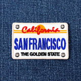 An embroidered patch designed like a California license plate reading 'SAN FRANCISCO' and 'THE GOLDEN STATE', placed on a blue denim background.