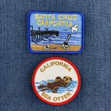 Two embroidered patches displayed on a blue denim fabric background. The top rectangular patch has a blue border and reads 'SANTA CRUZ CALIFORNIA' above a beach and pier scene. The bottom patch displays a sea otter with the words 'CALIFORNIA SEA OTTER'