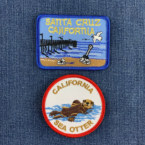 Two embroidered patches displayed on a blue denim fabric background. The top rectangular patch has a blue border and reads 'SANTA CRUZ CALIFORNIA' above a beach and pier scene. The bottom patch displays a sea otter with the words 'CALIFORNIA SEA OTTER'