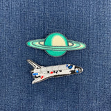 Two embroidered patches on blue denim: a green and cream planet with rings above and a white space shuttle with blue and red details below.