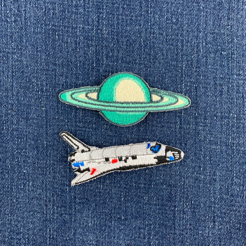 Two embroidered patches on blue denim: a green and cream planet with rings above and a white space shuttle with blue and red details below.