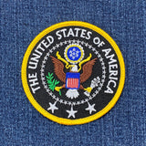 Round patch depicting the Great Seal of the United States, featuring a bald eagle, shield, olive branch, and arrows. Yellow text reads: "THE UNITED STATES OF AMERICA."