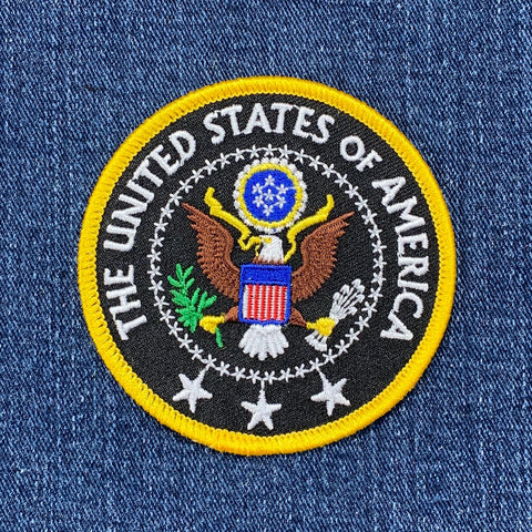 Round patch depicting the Great Seal of the United States, featuring a bald eagle, shield, olive branch, and arrows. Yellow text reads: "THE UNITED STATES OF AMERICA."