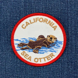 A circular embroidered patch featuring a California sea otter swimming in blue water, with 'CALIFORNIA SEA OTTER' embroidered in tan around the border, shown placed on blue denim fabric.