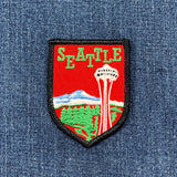 A shield-shaped embroidered patch featuring the word 'SEATTLE' in green on a red background, with a white space needle tower, blue mountain, and green trees, placed on blue denim fabric.