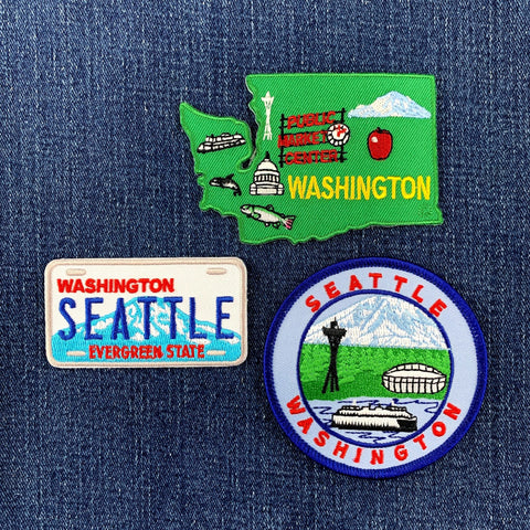 Three embroidered patches on a denim background: Washington state map, Seattle license plate, and Seattle cityscape.