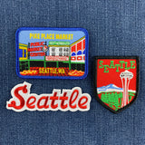 Three embroidered Seattle patches displayed on a blue denim background: a rectangular Pike Place Market patch and a shield-shaped Seattle patch below it, with a red-and-white embroidered 'Seattle' script patch on the bottom left.