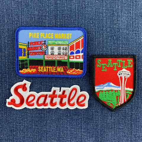 Three embroidered Seattle patches displayed on a blue denim background: a rectangular Pike Place Market patch and a shield-shaped Seattle patch below it, with a red-and-white embroidered 'Seattle' script patch on the bottom left.