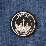 Circular embroidered patch with a black background and white border, featuring the Seattle skyline, including the Space Needle, outlined in white, and the words 'SEATTLE' and 'WASHINGTON' stitched around the top and bottom.