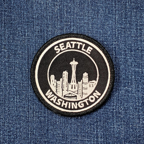 Circular embroidered patch with a black background and white border, featuring the Seattle skyline, including the Space Needle, outlined in white, and the words 'SEATTLE' and 'WASHINGTON' stitched around the top and bottom.