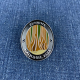 An oval enamel Walking Stick Medallion featuring a design of Sequoia National Park with stylized tree silhouettes in gold, green, and white on a black background, displayed on a blue denim surface.