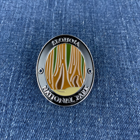An oval enamel Walking Stick Medallion featuring a design of Sequoia National Park with stylized tree silhouettes in gold, green, and white on a black background, displayed on a blue denim surface.