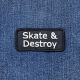 A rectangular black embroidered patch with white text reading 'Skate & Destroy' displayed on a blue denim fabric background.