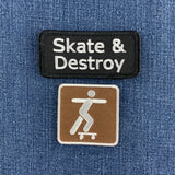 Two embroidered patches displayed on a blue denim fabric background. The top patch is black with white text reading 'Skate & Destroy'. The bottom patch is a brown and white skatepark sign.
